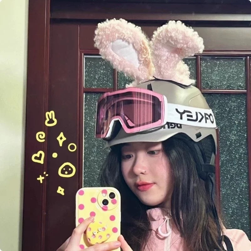 Person wearing a helmet with bunny ears and pink goggles, holding a phone with a yellow case.